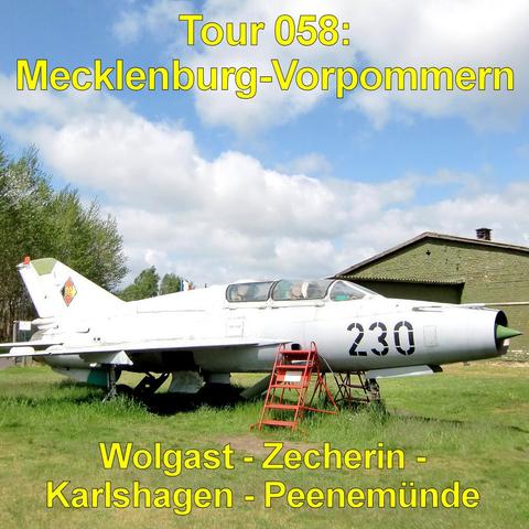 Ausgestelltes Militärflugzeug in grauer Lackierung steht auf einer Rasenfläche. Das Flugzeug besitzt eine spitze Nase, schmale Tragflächen und ein geschlossenes Cockpit mit transparenter Haube. Unter dem Rumpf sind Stützen angebracht. Eine rote Metalltreppe führt seitlich zum Cockpit hinauf. Auf dem Rumpf ist eine große dunkle Kennzeichnung angebracht. Im Hintergrund steht eine langgestreckte Halle mit grün gestrichener Fassade und leicht geneigtem Dach. Der Himmel ist blau mit großen, weißen W…