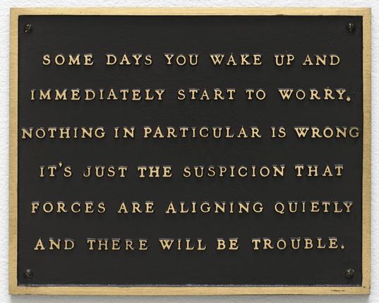 A rectangular bronze-style plaque with a dark, textured black background and a thin, metallic gold border. The plaque is secured with a screw in each of its four corners. Raised, all-caps gold lettering stretches across six lines, reading:

"SOME DAYS YOU WAKE UP AND
IMMEDIATELY START TO WORRY.
NOTHING IN PARTICULAR IS WRONG
IT’S JUST THE SUSPICION THAT
FORCES ARE ALIGNING QUIETLY
AND THERE WILL BE TROUBLE."