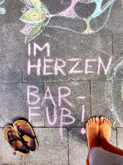 An overhead view of large concrete pavers where a message and drawings are rendered in pastel chalk art. The primary message is written centrally in pink block letters across two lines, reading "IM HERZEN BARFUß!" In the top left corner, there is a large, multicolored stylized chalk drawing of a lotus flower in teal, pink, and yellow. To the left of the text, a pair of worn brown leather strap sandals are placed on the paver. On the right, a pair of bare feet, appearing tan, are planted firmly …