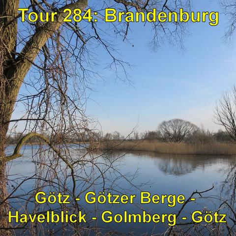 Ein ruhiger Flusslauf im Frühling. Die Bäume tragen keine Blätter. Die kahlen Äste spiegeln sich im stillen Wasser. Am Ufer stehen Schilf und niedrigere Vegetation. Der Himmel ist teilweise bewölkt, aber in einigen Bereichen blau und klar. Das Licht ist mild an einem sonnigen, aber kühlen Tag. Die Perspektive ist vom Ufer aus, mit Blick entlang des Flusses, umrahmt von den kahlen Ästen zweier großer Bäume im Vordergrund.