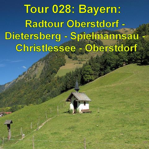 Eine kleine, weiße Kapelle mit schwarzem Dach steht auf einer grünen Wiese, die leicht an einem Berghang liegt. Im Hintergrund erstrecken sich bewaldete Hügel und Berge unter einem klaren blauen Himmel. Ein schmaler Weg führt zur Kapelle, entlang eines Zauns, der die Wiese umgibt. Rechts oben ist der Wald dichter, während links die Landschaft in weitere Täler übergeht.