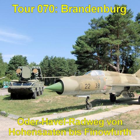 Ein ausgemustertes Militärflugzeug steht auf einem betonierten Streifen in einem Freigelände. Der Jet ist sandfarben lackiert, mit spitzer, grün gefärbter Nase, schmalem Rumpf und nach hinten gepfeilten Tragflächen. Das Cockpit ist geschlossen, das Fahrwerk ausgefahren, die Reifen stehen auf schmalen Betonplatten. Die Oberfläche wirkt stellenweise matt und leicht verwittert. Hinter dem Flugzeug befindet sich ein militärisches Fahrzeug mit großen Rädern und aufgerichteten Raketen- oder Radareinh…