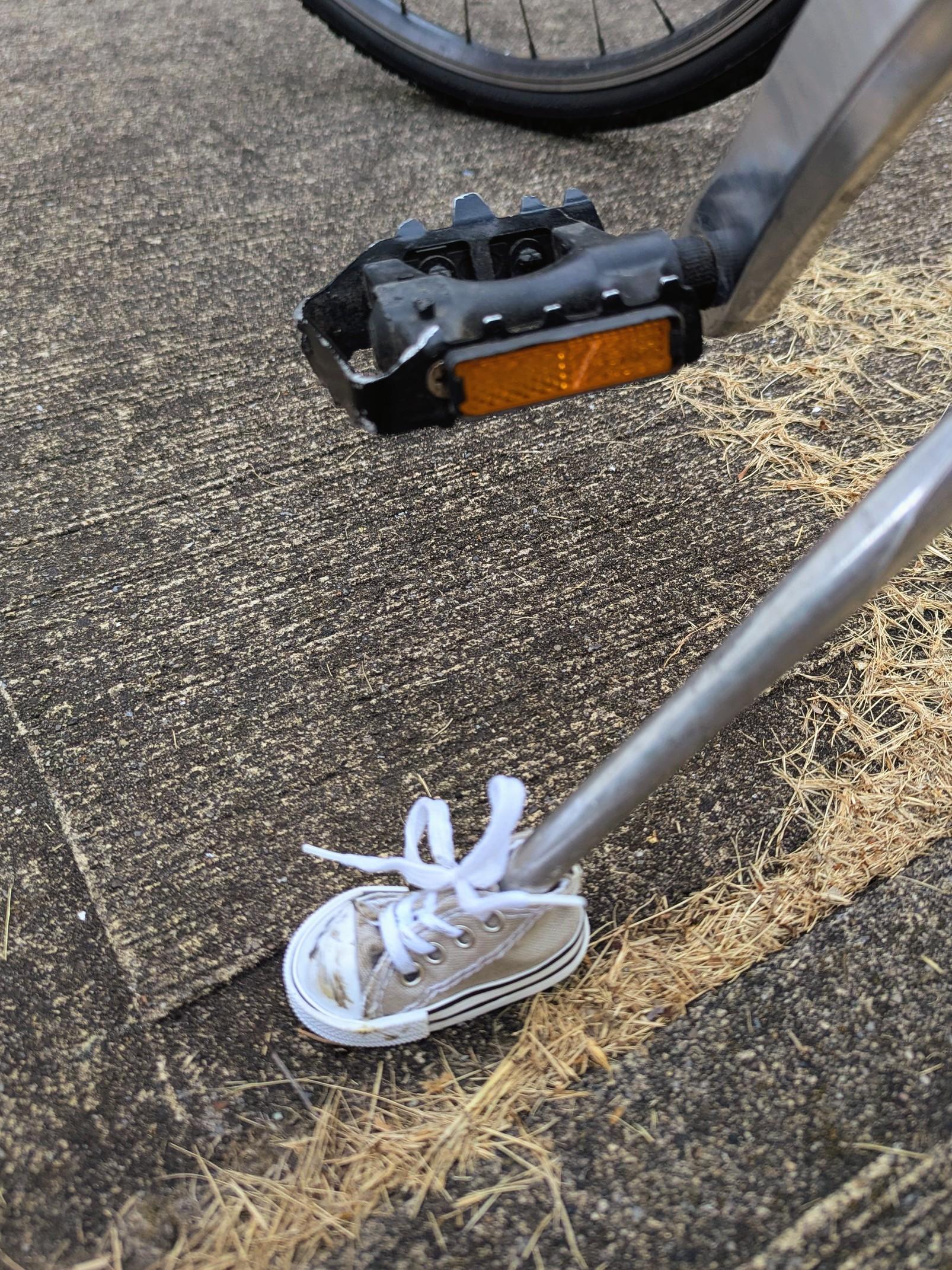 Looking at a concrete driveway with some dried grass. A tiny grey sneaker with white laces is at the end of an aluminum rod that extends up and out of frame. A bike pedal, black and with an orange reflector is in frame and at top of frame is the small arc of the front tyre and spokes of the bike