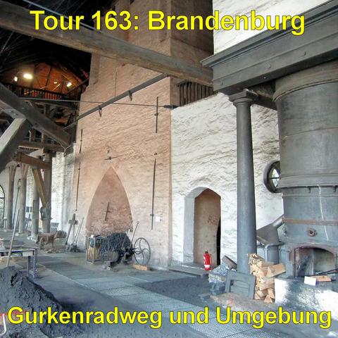 Großer Innenraum mit hohen, weiß verputzten Wänden und sichtbaren Holzbalken unter einem gewölbten Dach. Entlang der Wand stehen alte Werkzeuge und Metallgeräte, darunter lange Stangen, Kreuze aus Metall und eine mechanische Vorrichtung mit großen Rädern. In der rechten Bildhälfte befindet sich ein massiver, zylindrischer Ofen aus Metall mit einer geöffneten Feuerkammer. Daneben liegt gestapeltes Brennholz. Auf dem Boden sind dunkle Kohlereste verteilt, teilweise in einer länglichen Aufschüttun…