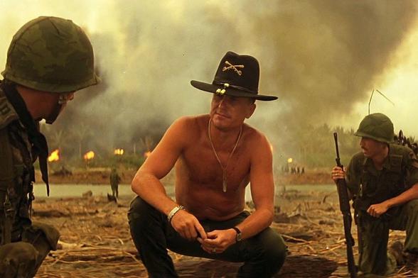 A still from the film Apocalypse Now showing a shirtless U.S. Army officer wearing a black cavalry hat and dog tags crouching between two armed soldiers on a smoke-filled battlefield. Fires burn in the background near a river, with additional troops visible in the distance.