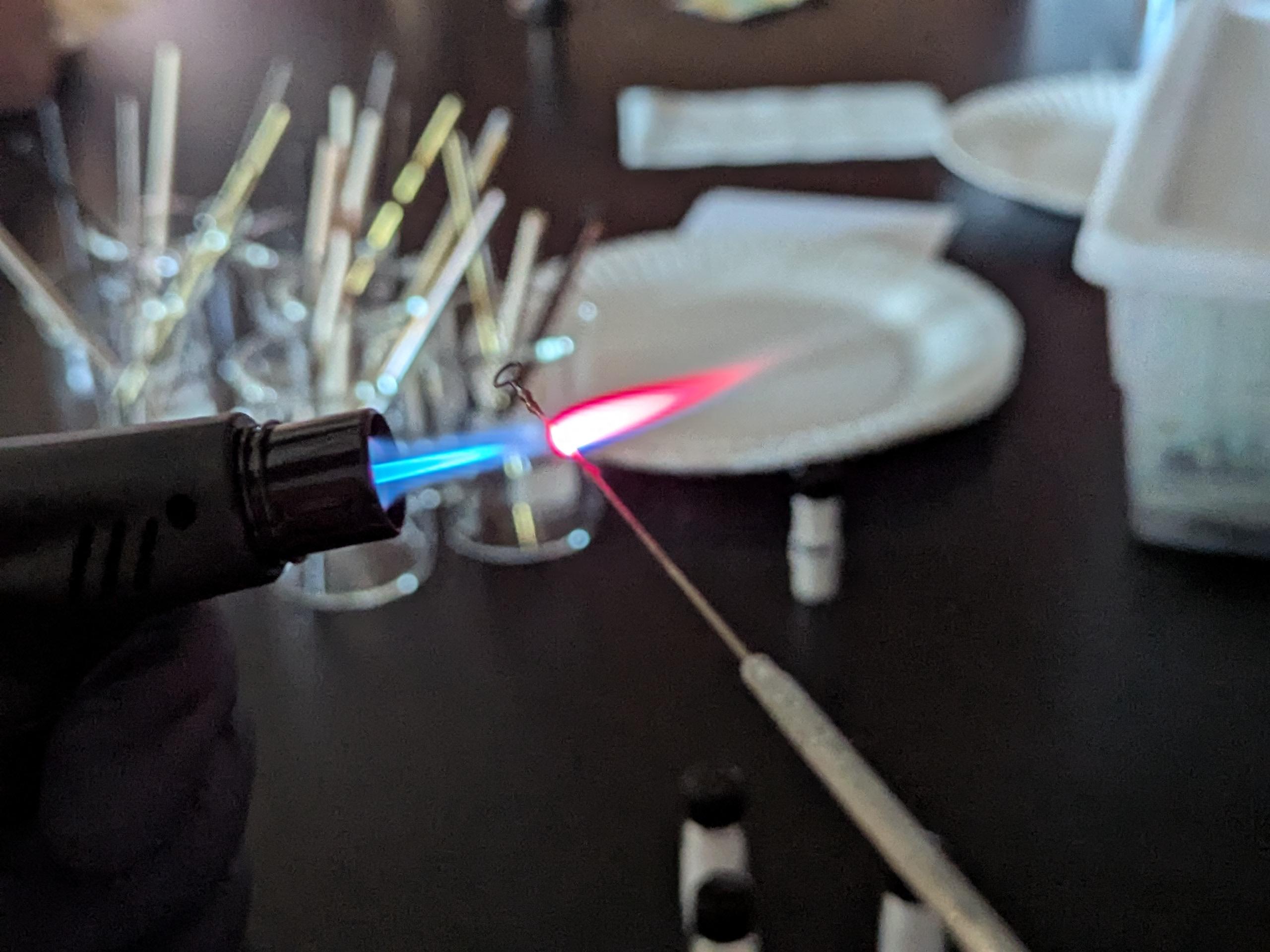 A small butane torch with thin blue flame and a thin iron rod with loop held in front of the blue flame causing the jet to be bright red