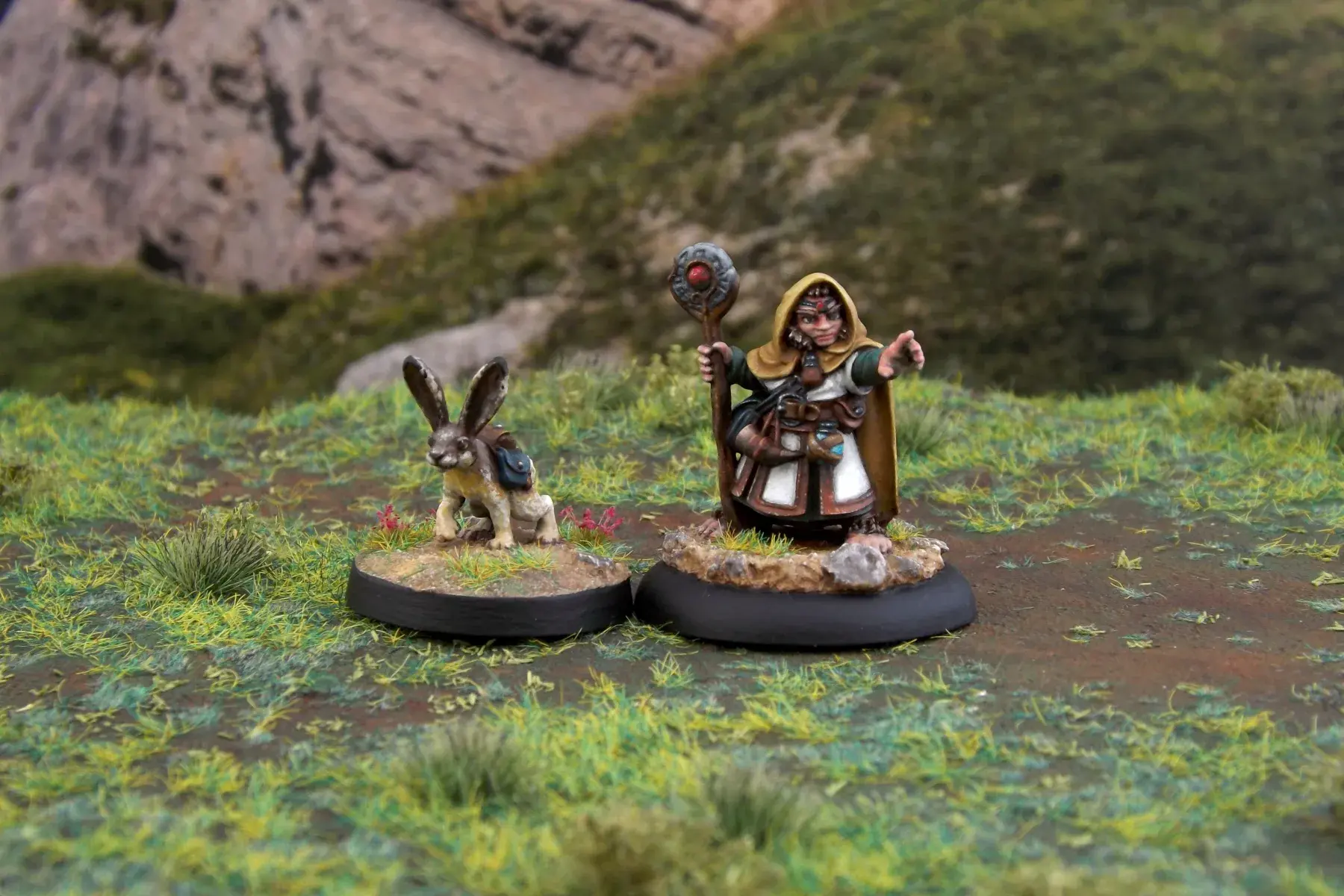 A painted miniature of a halfling druid with his hare companion.  The hare, on the left, is brown and gray and is wearing several packs on its back.  The druid, on the right, has a hooded ochre cloak and a wooden staff with a stone headpiece.  It is photographed on grassy terrain.