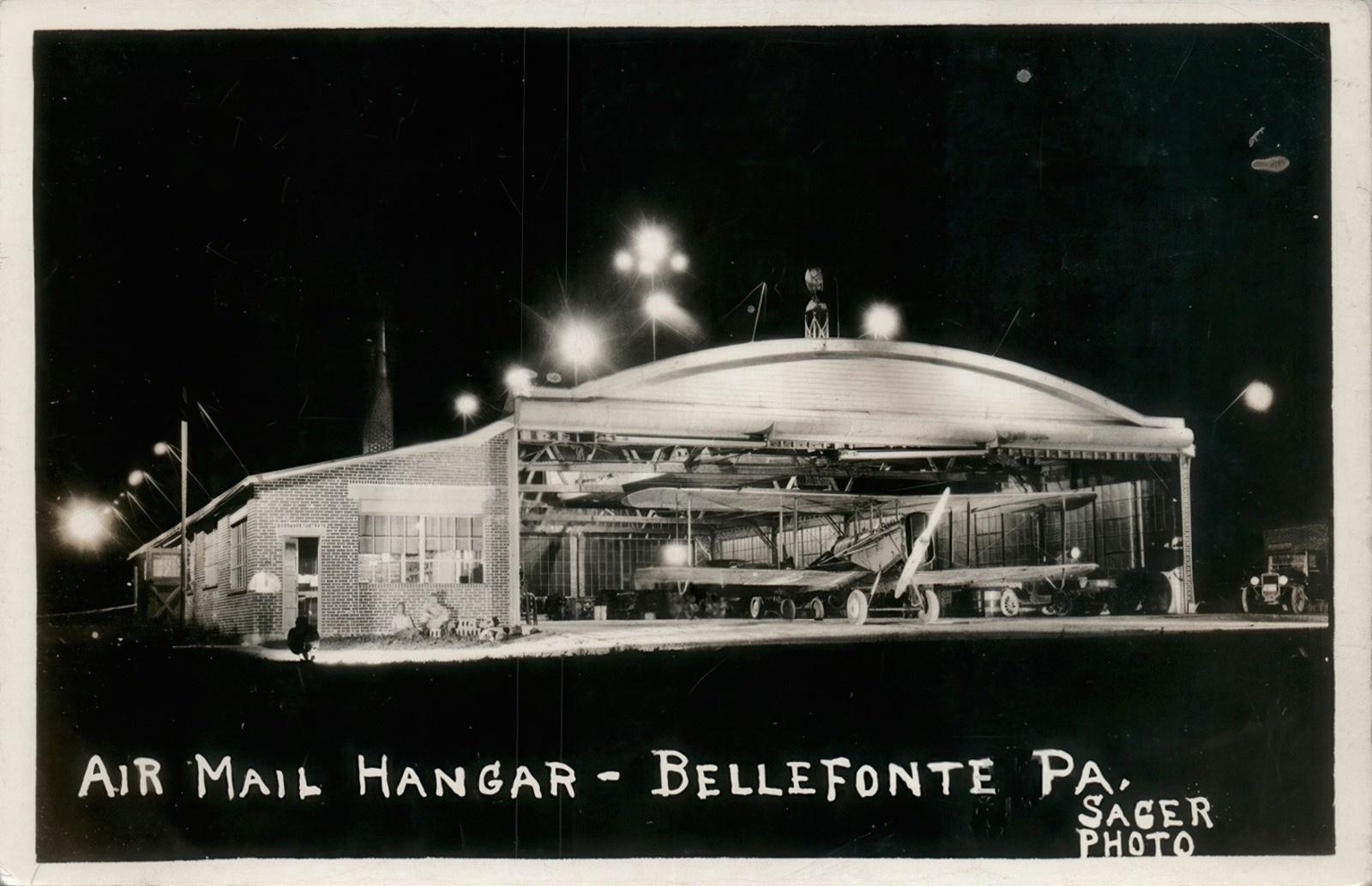 Black-and-white real photo postcard showing the illuminated Air Mail Hangar at the Bellefonte Airport in Bellefonte, Pennsylvania, at night. The large arched hangar stands with its doors fully open, revealing several early single-engine airplanes parked inside under bright overhead lights. A one-story brick office building adjoins the hangar on the left, while a vintage automobile is visible near the right edge of the image. The dark night sky contrasts with the brightly lit airfield, emphasizing the airport's role in early airmail operations. Hand-lettered text along the bottom reads, “Air Mail Hangar – Bellefonte Pa.,” with the photographer's credit, “Sager Photo,” in the lower-right corner.