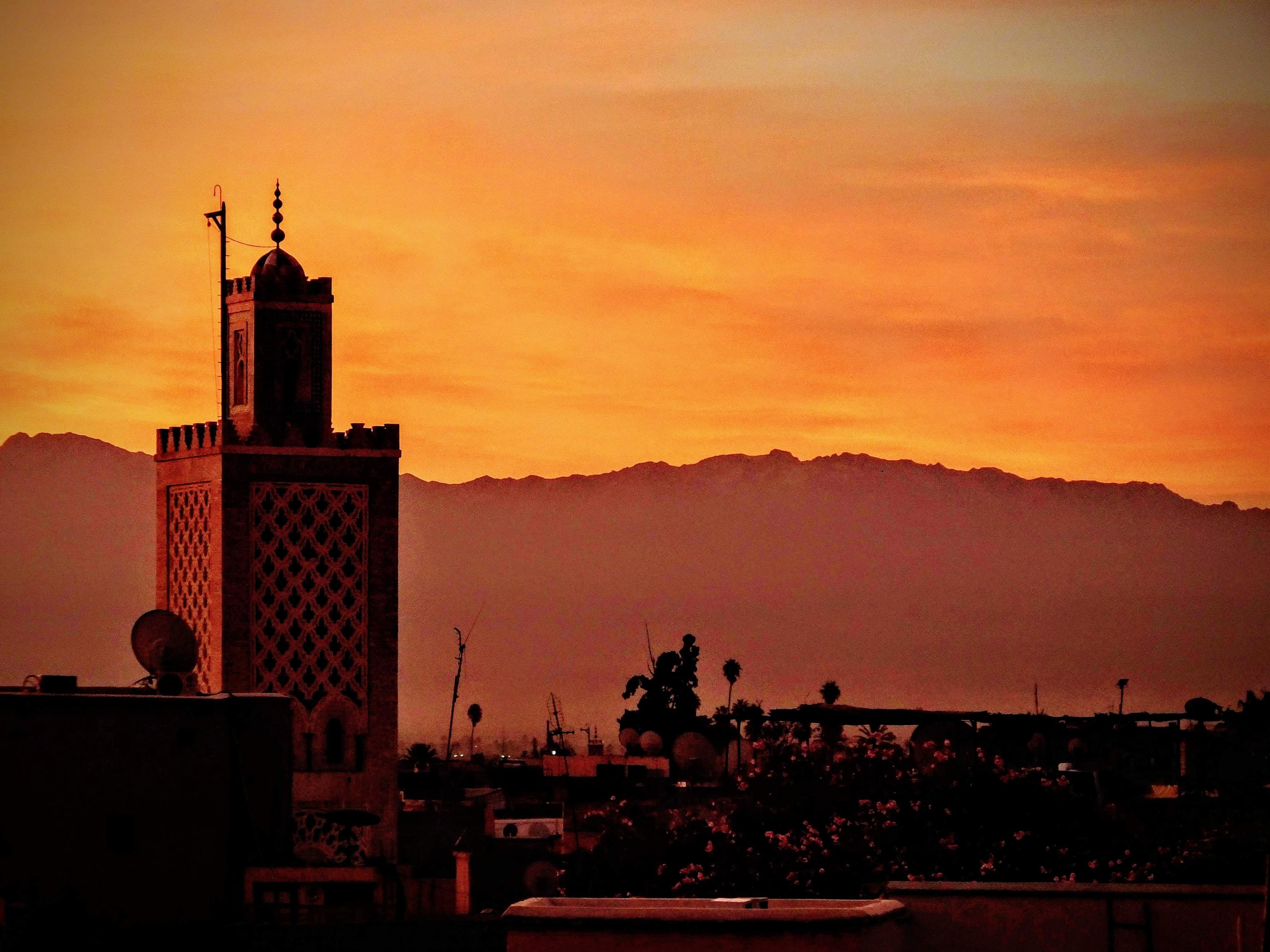 Yellow orange sky with the silouhette of mountains in the background across the middle of the frame. The tower of the Koutoubia mosque to the left in the red light of sunrise dawn