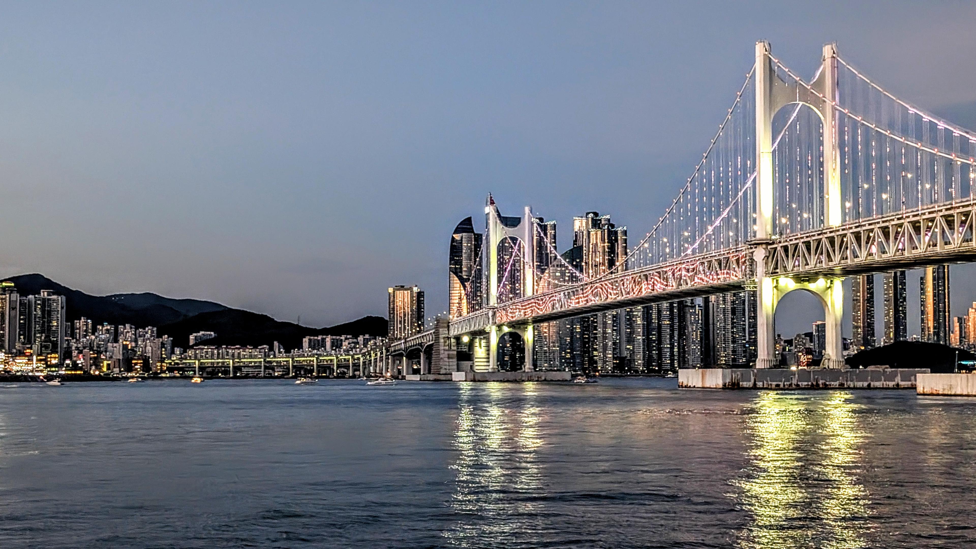 Colour photo of a bridge illuminated during sunset. In the background is the city with high rise buildings. Taken from a cruise boat on a trip around the harbour.