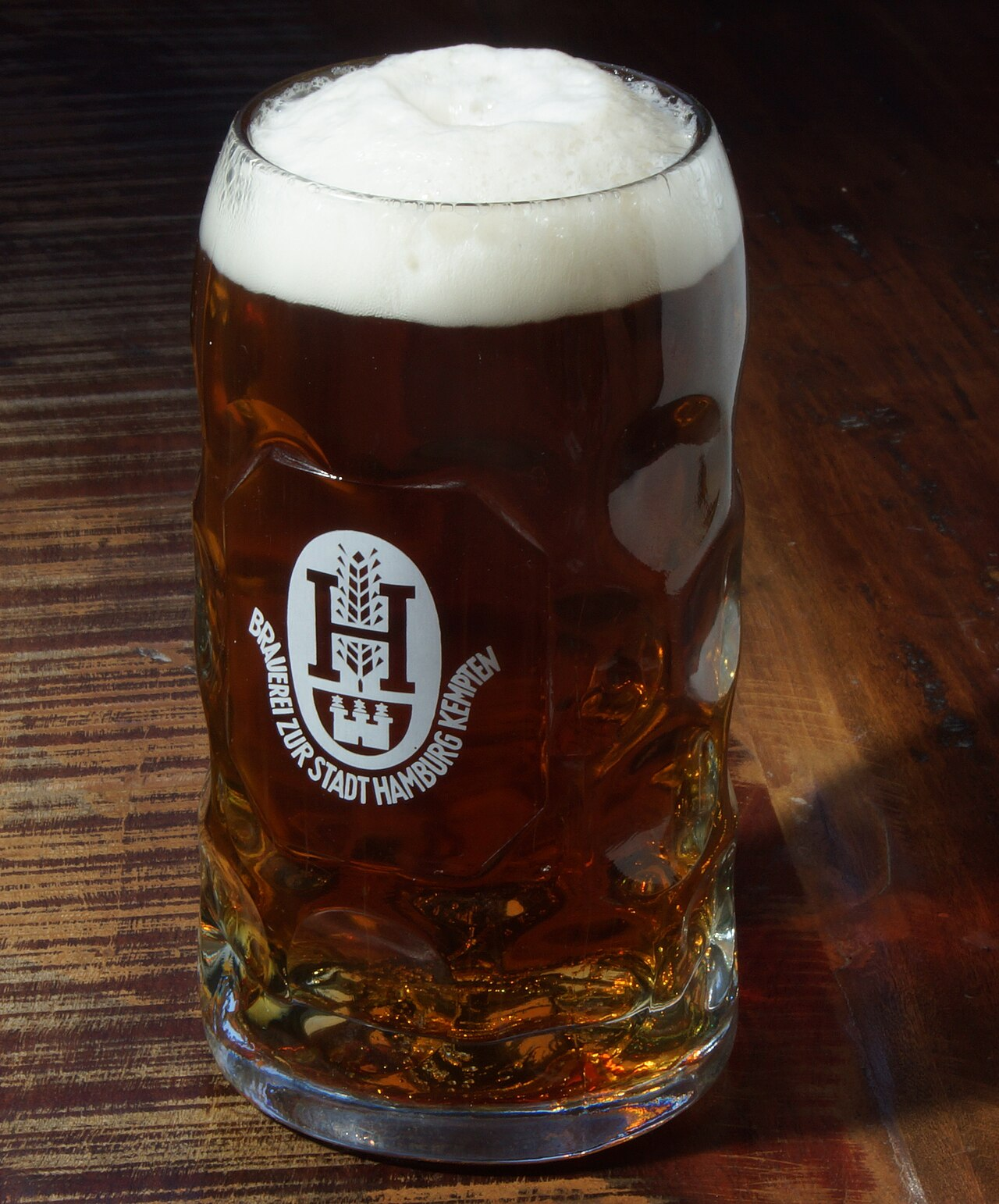 Ein großer, voller Maßkrug Bier mit goldbrauner Farbe und dicker weißer Schaumkrone auf einem Holztisch.