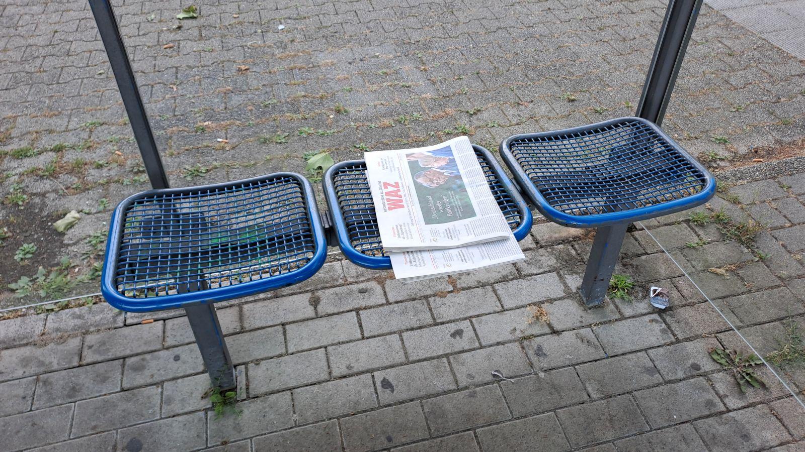 Dreigeteilte Sitzbank in einem Haltestellenhäuschen. Auf dem mittleren Sitz liegt eine ordentlich zusammengefaltete Tageszeitung.