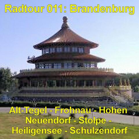 Ein großes, rundes Gebäude mit mehrstöckigem, pagodenartigem Dach erhebt sich auf einer erhöhten Plattform. Das Dach ist dunkelrot mit geschwungenen Linien und einer goldfarbenen Spitze. Mehrere Etagen mit umlaufenden Balkonen und roten Säulen sind übereinander angeordnet. Eine breite Treppe führt zur oberen Ebene hinauf. Vor dem Bauwerk erstreckt sich eine gepflegte Rasenfläche mit kleinen Sträuchern und symmetrisch angelegten Wegen. Im Hintergrund stehen dichte grüne Bäume unter einem klaren …