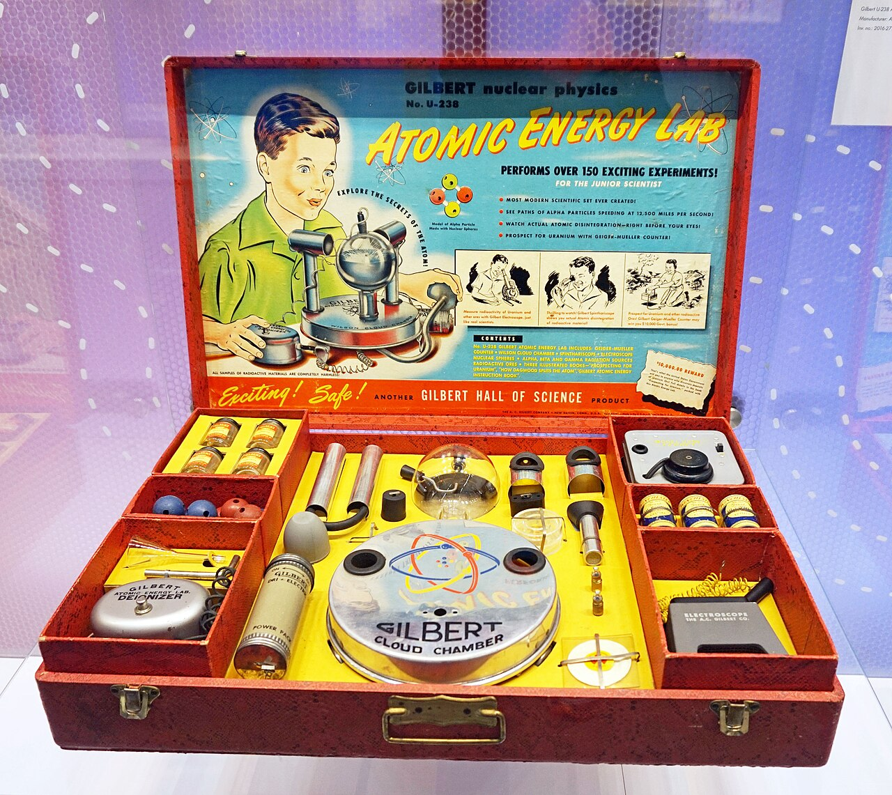 Ein geöffneter Experimentierkasten aus den 1950ern: „Gilbert Atomic Energy Lab U-238“. Im roten Koffer liegen Messgeräte, Döschen, eine Nebelkammer und Anleitungen; im Deckel ein gezeichneter Junge.