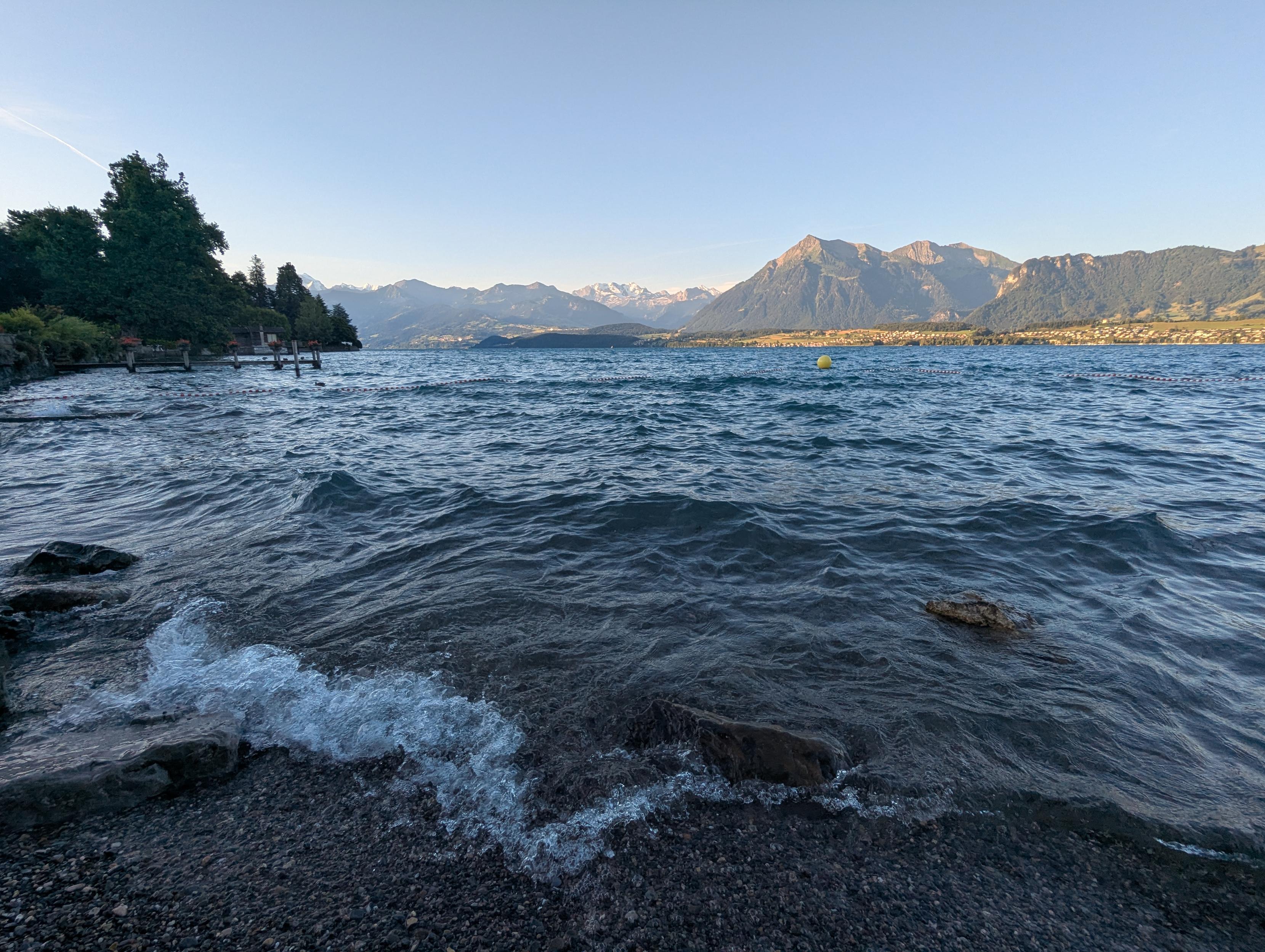 The shore of a lake in the shadow, some small waves. Mountains in the background, brightly lit by the sun.