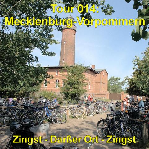 Backsteingebäude mit rundem, hohen Turm steht inmitten von Bäumen. Der Turm hat eine leicht verjüngte Form und trägt eine kleine Plattform mit Geländer. Vor dem Gebäude befindet sich ein großer Fahrradparkplatz mit zahlreichen abgestellten Fahrrädern. Mehrere Personen bewegen sich zwischen den Rädern oder stehen daneben. Der Boden ist sandig, umgeben von Grünflächen und Laubbäumen. Das Gebäude besitzt helle Fensterbögen und ein flach geneigtes Dach. Die Szene wirkt wie ein beliebtes Ausflugszie…