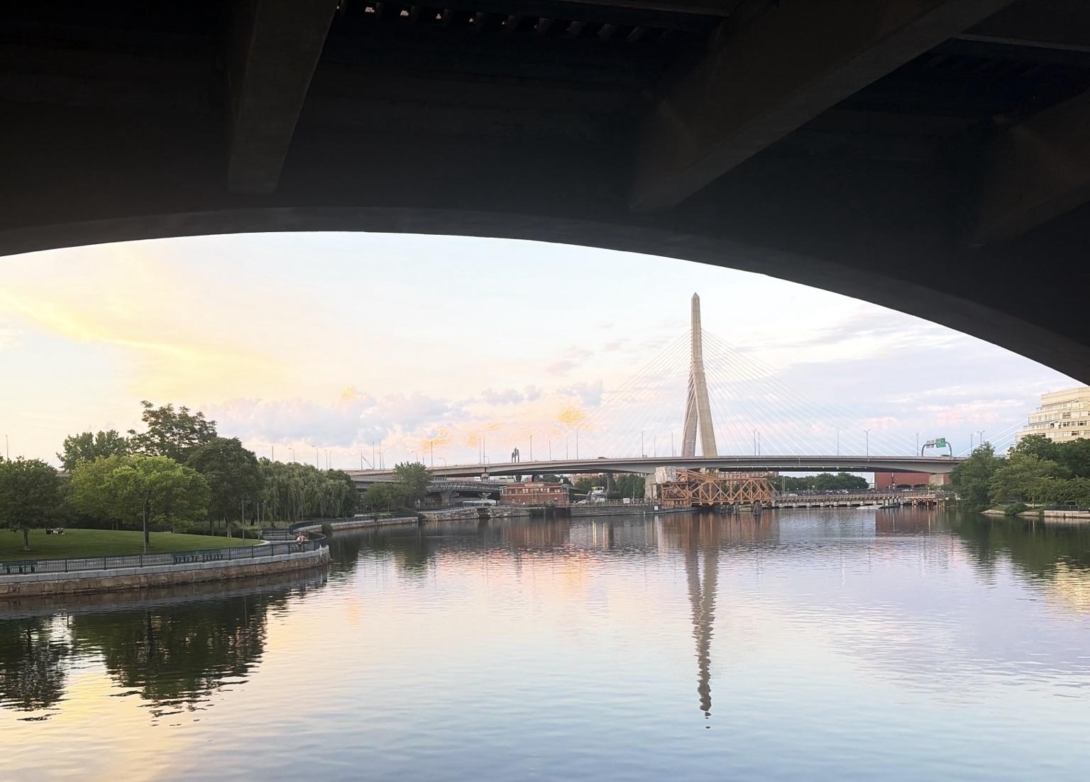 A sunset view of a bridge with a prominent tall structure, reflecting in the calm water below. The scene is framed by an arch from an overhanging structure, with greenery visible along the water's edge and a pastel sky above.