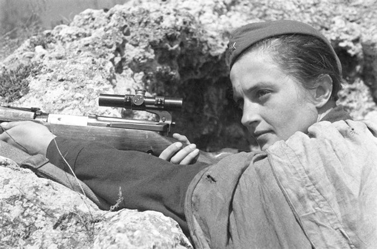 Black and white photo of Lyudmila Pavlichenko holding a rifle with optical site while standing in a trench facing to the left.  She is wearing a barracks military cap and appears to have shawl draped over her shoulders.