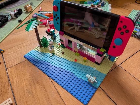 An eye-level photograph of a custom-built LEGO diorama of a detailed beach and hut scene, which is serving as a functional, decorative stand for a Nintendo Switch console. The scene is constructed on a rectangular LEGO baseplate, split between light blue water (the 'ocean') and tan sand (the 'beach'). The water has small, simple clear LEGO elements and a small pink buildable character. The pink and magenta-colored beach hut structure is centrally placed, supporting the black Nintendo Switch scr…