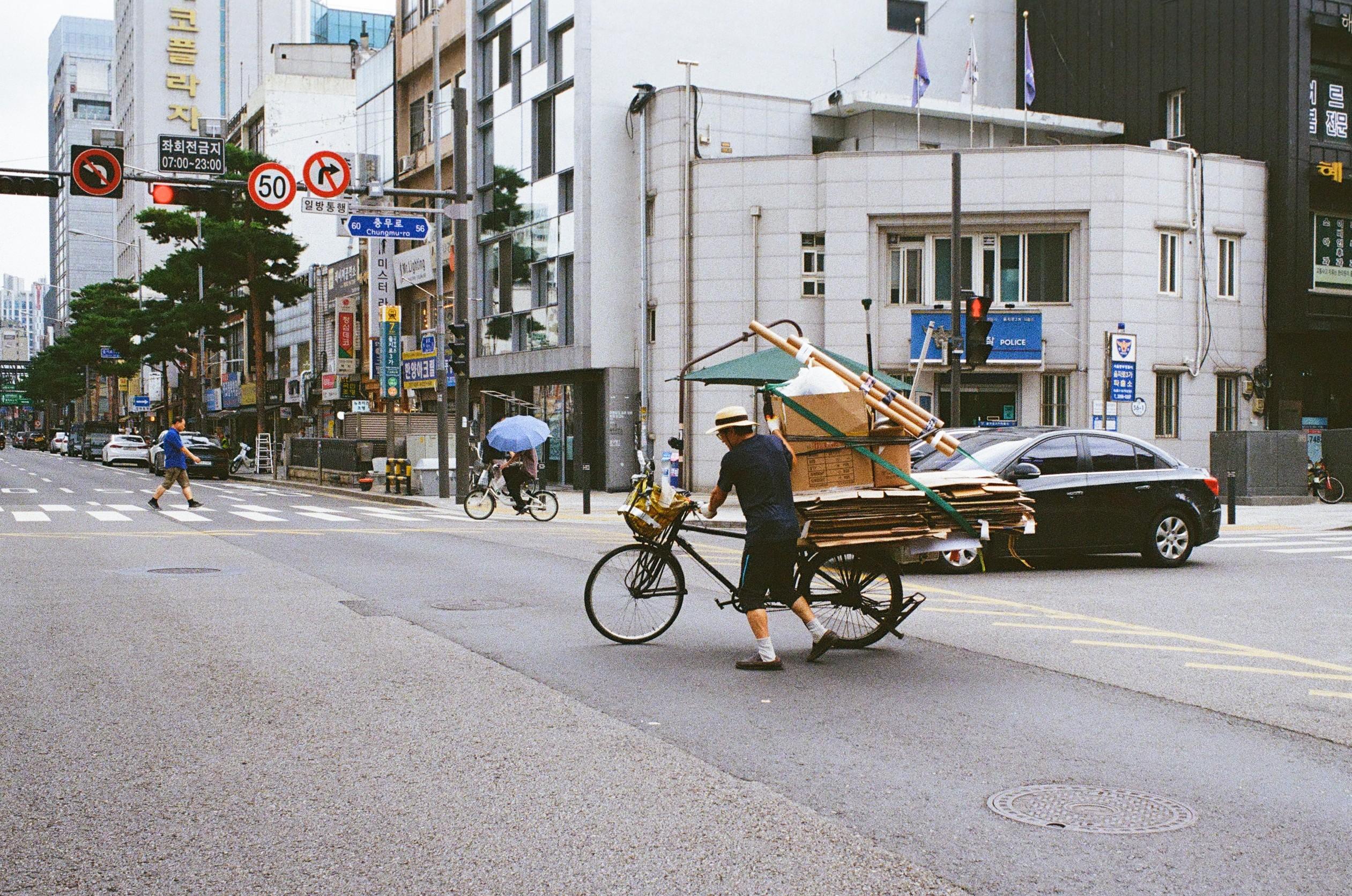 Colour 35mm photo, Pentax Spotmatic 500, Kodak Ultra Max 400, SMC Takumar 35mm lens. Developed at at lab in Seoul, scanned there by myself. Image shows a gentleman pushing a bicycle loaded with flattened cardboard, boxes and other objects. In the background a gentleman is walking a zebra crossing. It was a hot and humid day, but heavy rain and thunderstorms were due later in the afternoon.