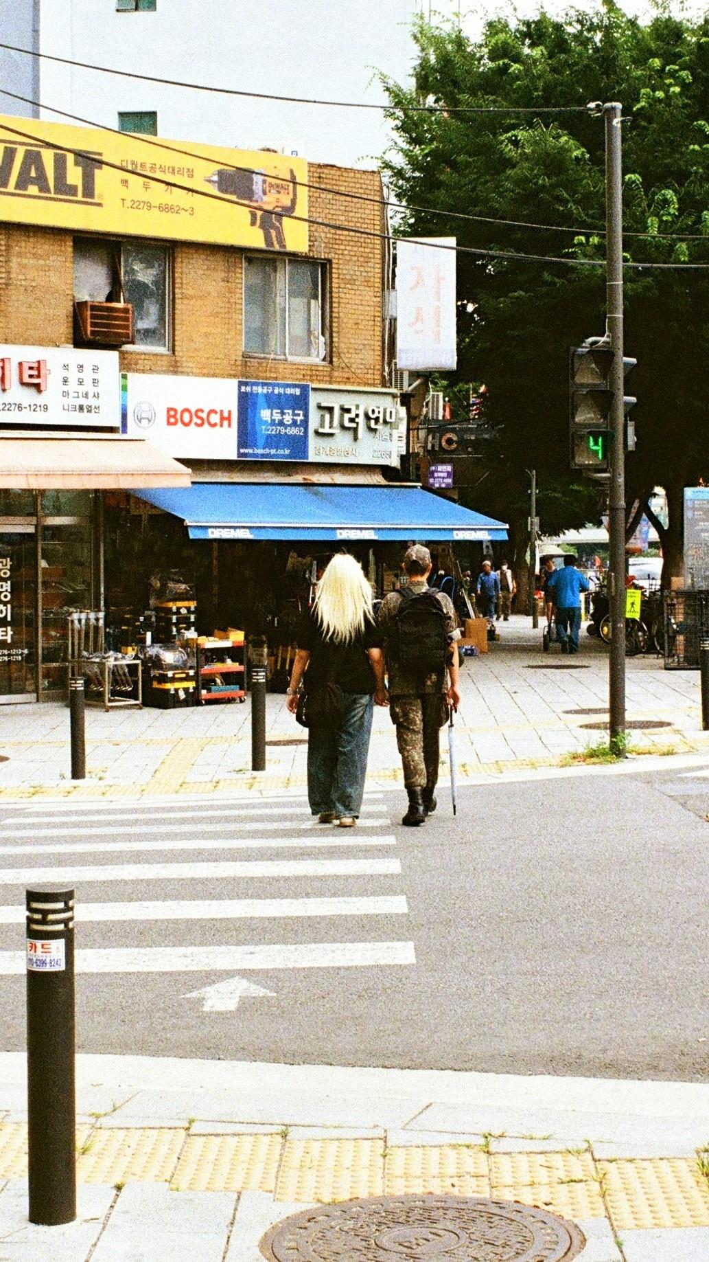 Colour 35mm photo, Pentax Spotmatic 500, Kodak Ultra Max 400, SMC Takumar 35mm lens. Developed at at lab in Seoul, scanned there by myself. Image shows a Korean soldier with his partner on a zebra crossing. He is holding her hand and carrying an umbrella. Seeing Korean soldiers in uniform (on leave) in the city was a very common sight.