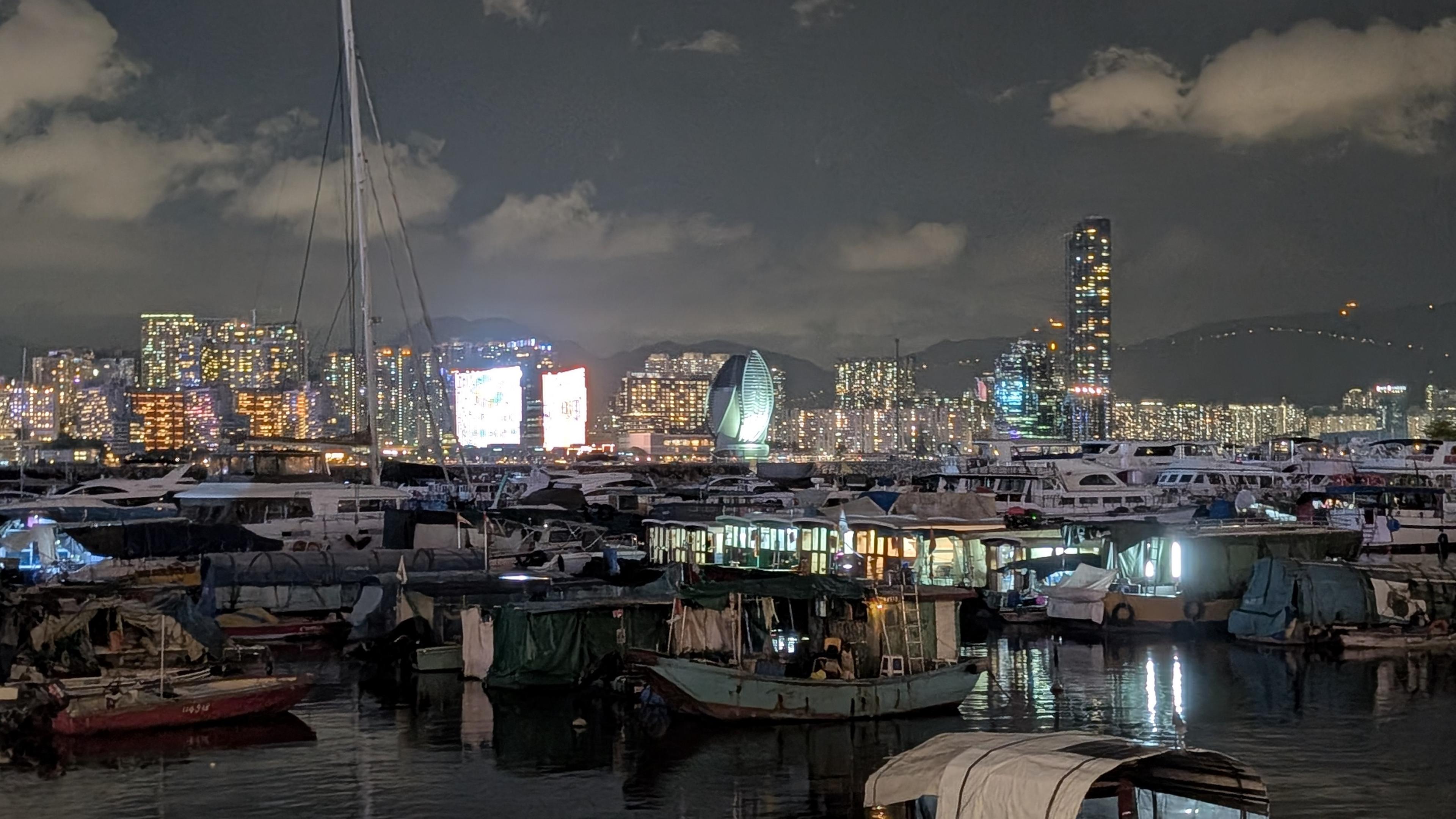 Colour photo of a view across the harbour, North Point, Hong Kong at night.
Image shows boats of all sizes moored in the harbour. In the background are a section of modern high rise buildings.