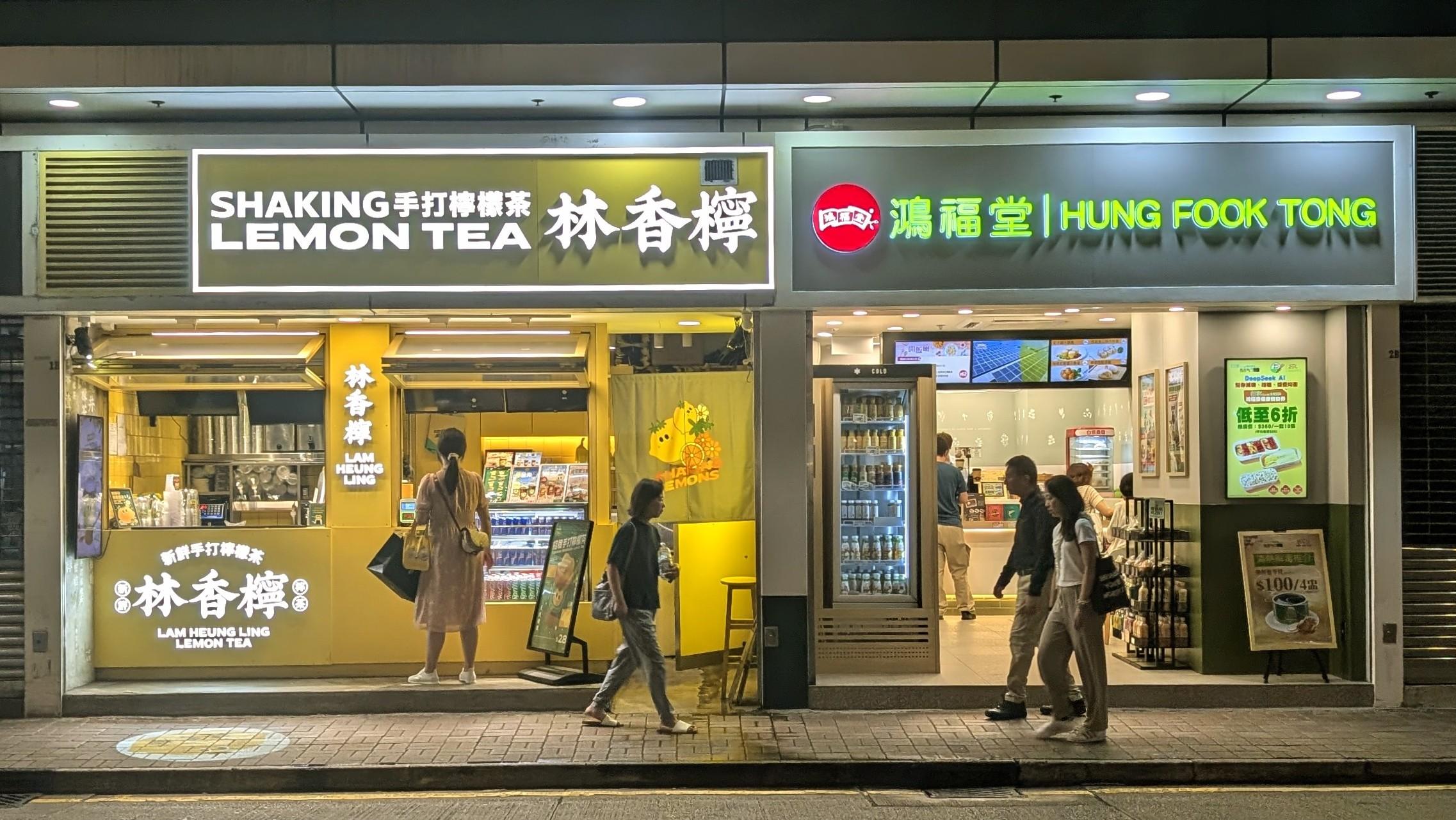 Colour photo of two shops in the North Point area of Hong Kong at night. Image shows people walking by and customers in the shops. One sells lemon tea as well as other cooling drinks required in the humidity, the other is a fast food takeaway.