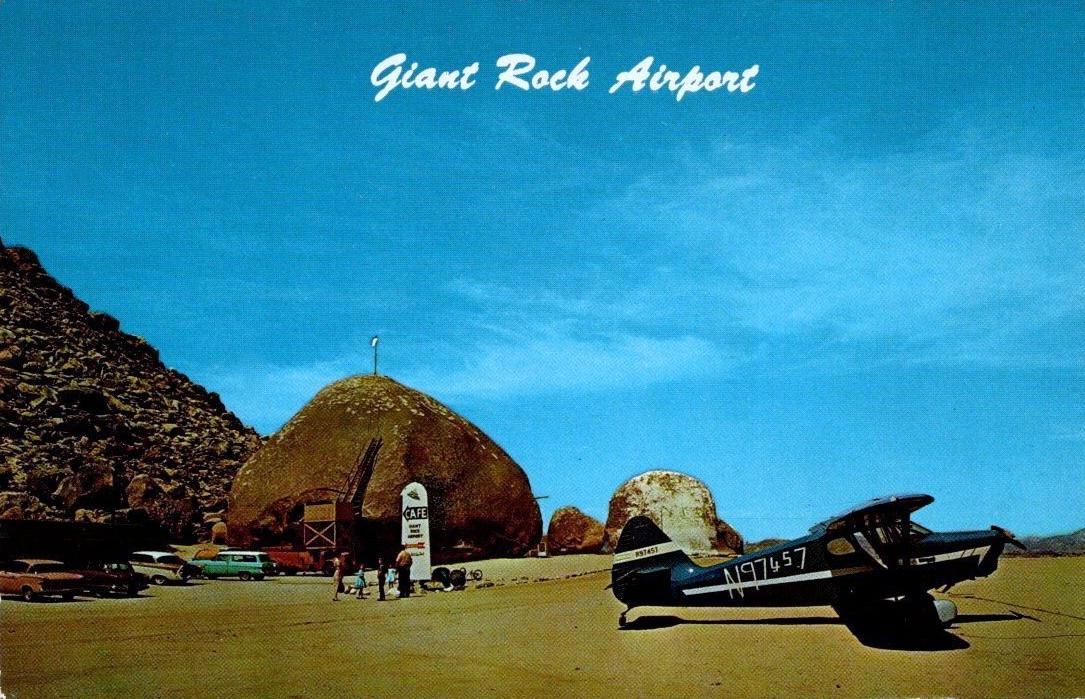 A vintage color postcard titled "Giant Rock Airport" shows one of the most unusual private airfields in the California desert. The scene is set beneath a brilliant blue sky streaked with wispy white clouds, with the vast, sandy desert stretching across the foreground.

Dominating the left side of the image is the enormous rounded granite boulder known as Giant Rock, one of the world's largest freestanding boulders. Nestled against its base is the airport's distinctive café and small visitor area, built directly into and around the massive rock. A wooden staircase climbs the face of the boulder toward its summit, where a small flagpole rises from the top. In front of the café stands a roadside-style sign reading "Cafe," while several visitors gather nearby, including adults and children exploring the attraction.

Parked along the base of the rock are several late-1950s or early-1960s automobiles in muted shades of green, tan, and white, reinforcing the postcard's mid-century era. The vehicles are lined up beside the café, suggesting both tourists and pilots visiting the remote desert destination.

The right half of the postcard features a dark-colored, high-wing, single-engine tailwheel airplane parked on the sandy airstrip. The aircraft is viewed in profile with its nose pointed to the right, its shadow stretching beneath it in the bright desert sun. The registration number N97457 is painted in large white characters along the fuselage, and the aircraft appears ready for departure from the informal desert landing field.

In the distance, low desert mountains form the horizon beneath the expansive sky, emphasizing the isolated setting of Giant Rock Airport. The postcard captures the unique combination of aviation, roadside tourism, and California desert culture that made Giant Rock a famous destination for private pilots, adventurers, and sightseers during the mid-twentieth century. The image conveys both the novelty of landing beside one of the world's largest boulders and the wide-open freedom associated with general aviation in the American Southwest.