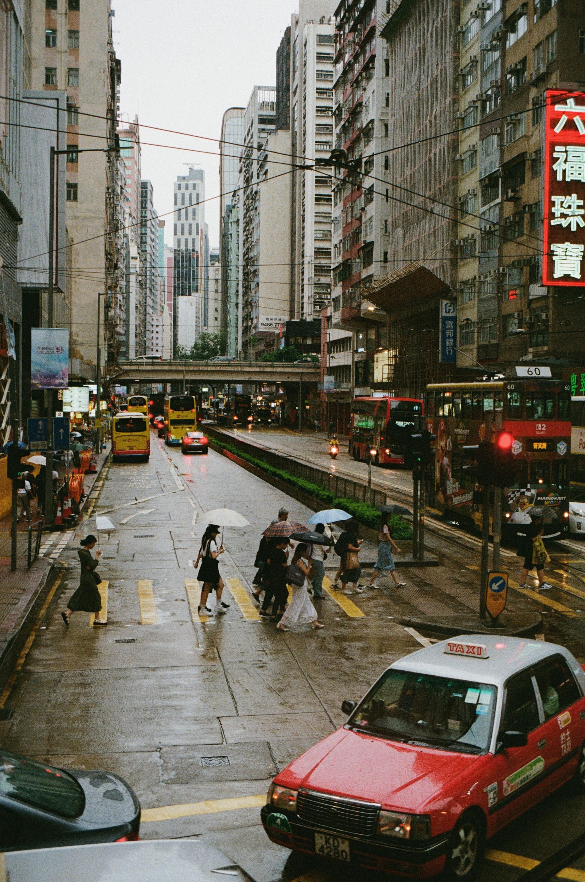 Colour photo of a street in Hong Kong during the rain. Many people are crossing the road some holding umbrellas. A bright red taxi is turning off the street. Buses are in the distance There is an overpass road above the street. The street is surrounded by high rise buildings.