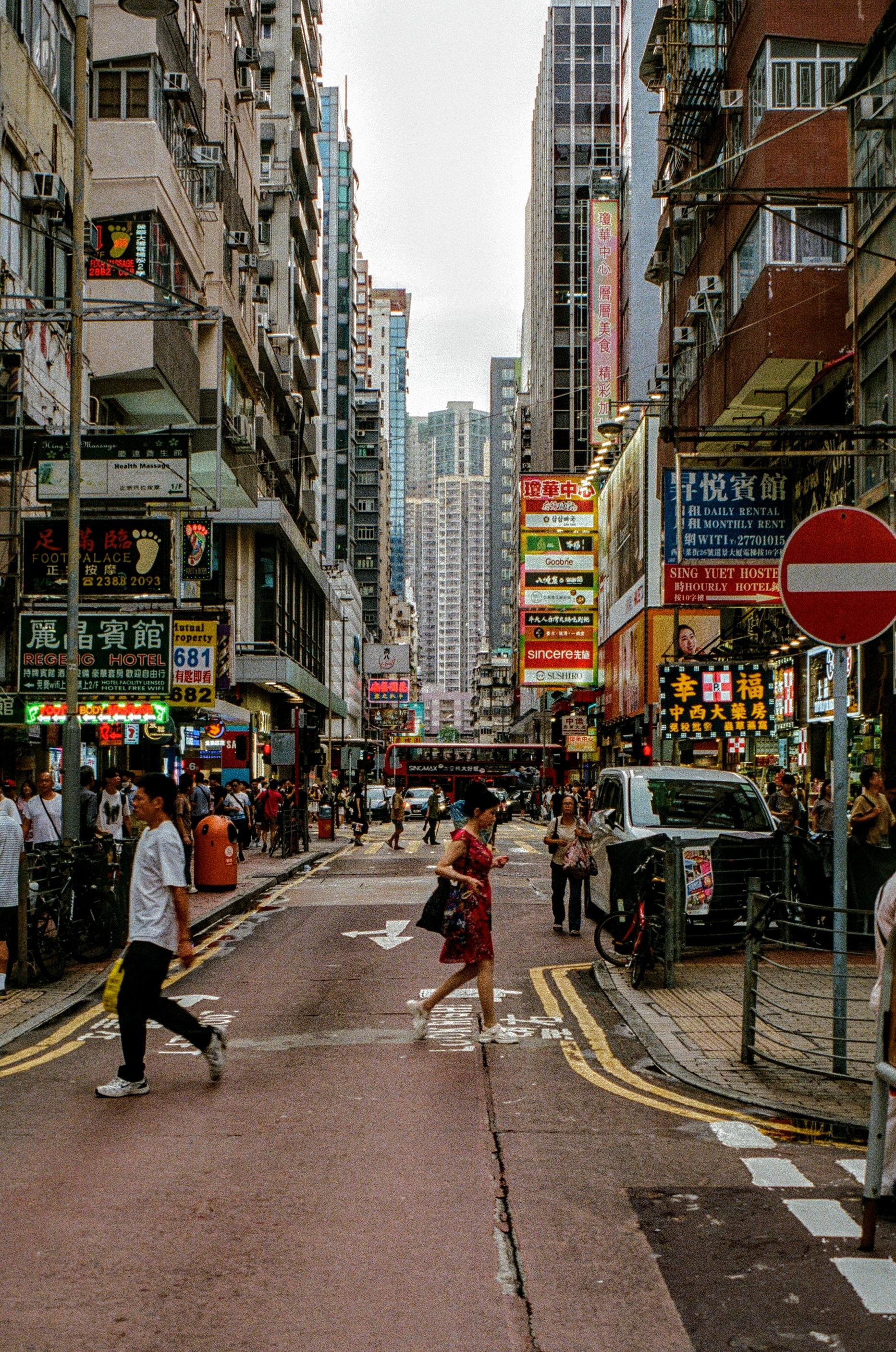 Colour photo of a street scene in Hong Kong. The rain has now passed but the skies are still grey and undecided. A young lady in a red dress is crossing the road while a gentleman in a white shirt is walking across the other side. The street is surrounded by high rise buildings, lots of advertising signs and lights are attached.