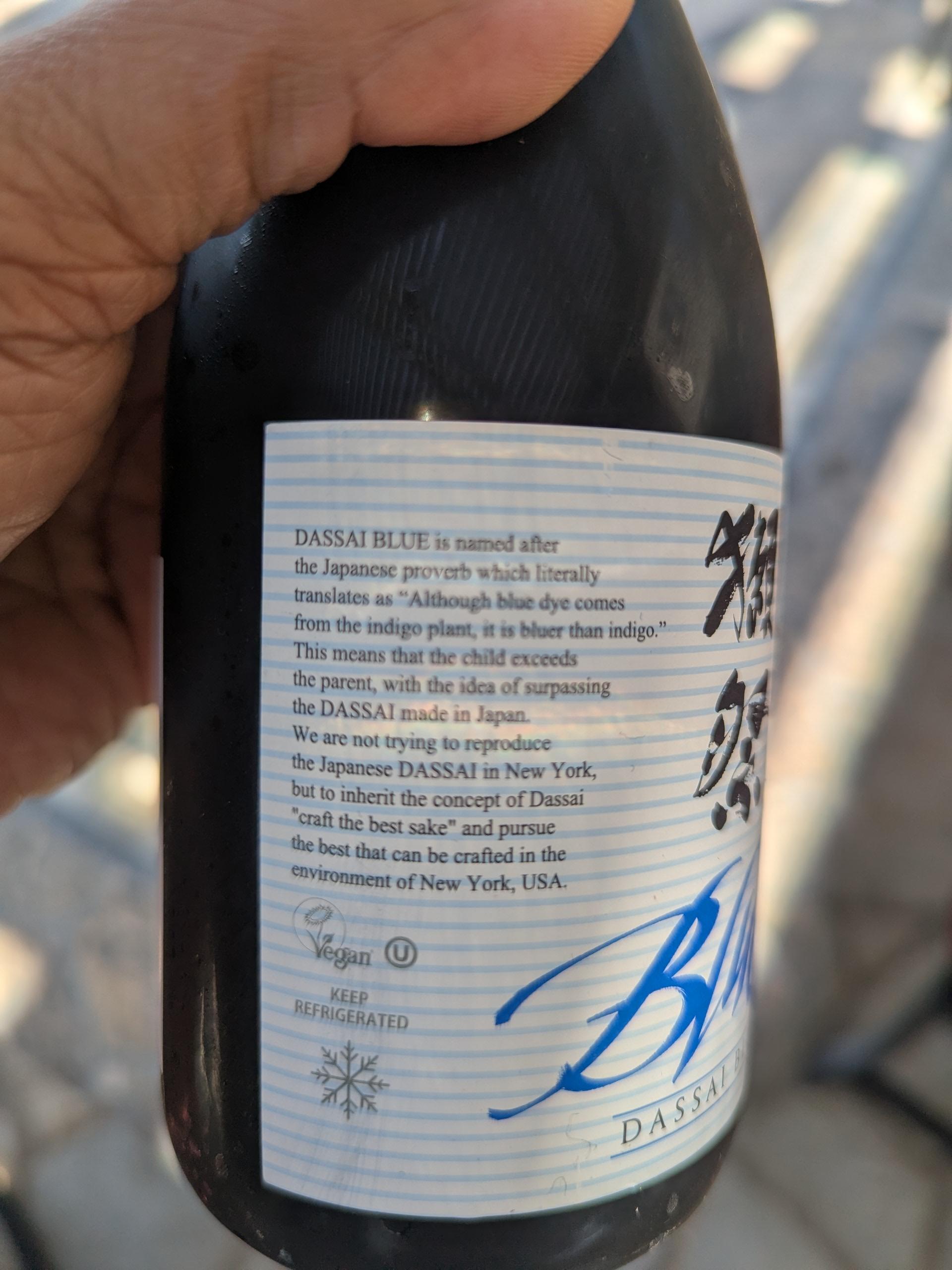 Holding the bottle to show the side of the white label on the dark bottle. 
Text reads
DASSAI BLUE is named after the Japanese proverb which literally translates as "Although blue dye comes from the indigo plant, it is bluer than indigo." This means that the child exceeds the parent, with the idea of surpassing the DASSAI made in Japan. We are not trying to reproduce the Japanese DASSAI in New York, but to inherit the concept of Dassai "craft the best sake" and pursue the best that can be crafted in the environment of New York, USA.
Vegan
KEEP REFRIGERATED