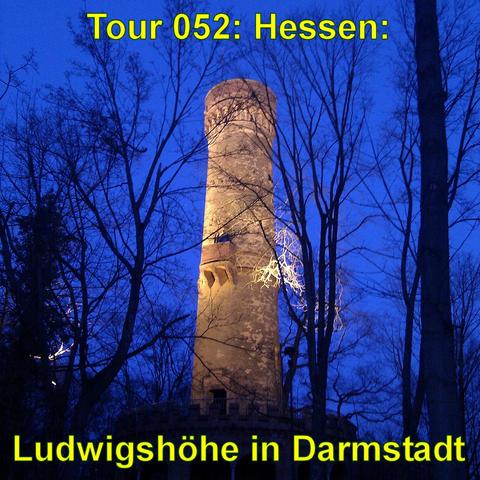 Ein beleuchteter Turm bei Dämmerung. Der Turm steht zwischen kahlen Bäumen und ragt mit seiner runden Struktur hervor. Seine Oberfläche ist von der Beleuchtung angestrahlt, was ihm einen warmen, leicht goldenen Ton verleiht. Die Szenerie wird durch das bläuliche Licht der frühen Nacht ergänzt, was einen Kontrast zwischen dem künstlichen Licht des Turms und dem natürlichen Licht des Himmels schafft. Die Bäume ohne Blätter deuten auf eine kältere Jahreszeit hin.