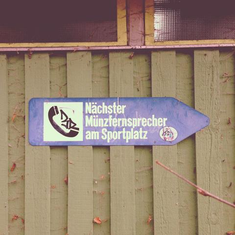 Ein blaues, pfeilförmiges Schild ist an einer Wand aus vertikalen grünen Holzbrettern angebracht. Auf der linken Seite des Schildes befindet sich ein weißes Symbol eines Telefonhörers und daneben steht der Text „Nächster Münzfernsprecher am Sportplatz“. An der Spitze des Pfeils ist eine kleine kreisförmige Grafik zu sehen. Die grünen Holzbretter im Hintergrund haben einige vertrocknete, braune Blätter an ihnen haften. Im oberen Bildrand ist ein Teil eines Fensterrahmens sichtbar.