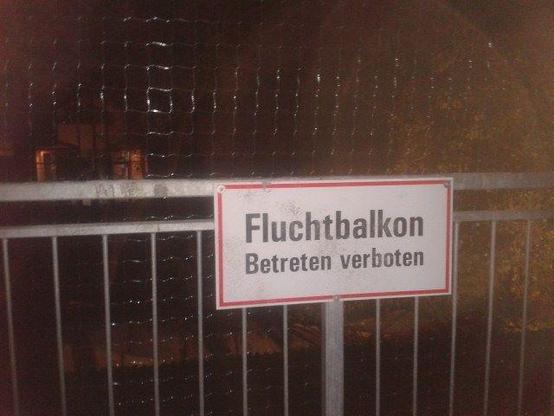 Ein Foto eines weißen Metallschilds mit einem roten Rand, das an einem grauen Metallgitterzaun befestigt ist. Auf dem Schild steht in schwarzer, serifenloser Schrift, zweizeilig: in der oberen Zeile 'Fluchtbalkon' (fettgedruckt und größer) und darunter 'Betreten verboten' (normal gedruckt, kleiner). Das Foto wurde bei Nacht aufgenommen, mit künstlichem Licht, das direkt auf das Schild fällt. Hinter dem Zaun ist ein weiteres Maschendrahtgitter zu sehen, und im dunklen Hintergrund sind verschwomm…