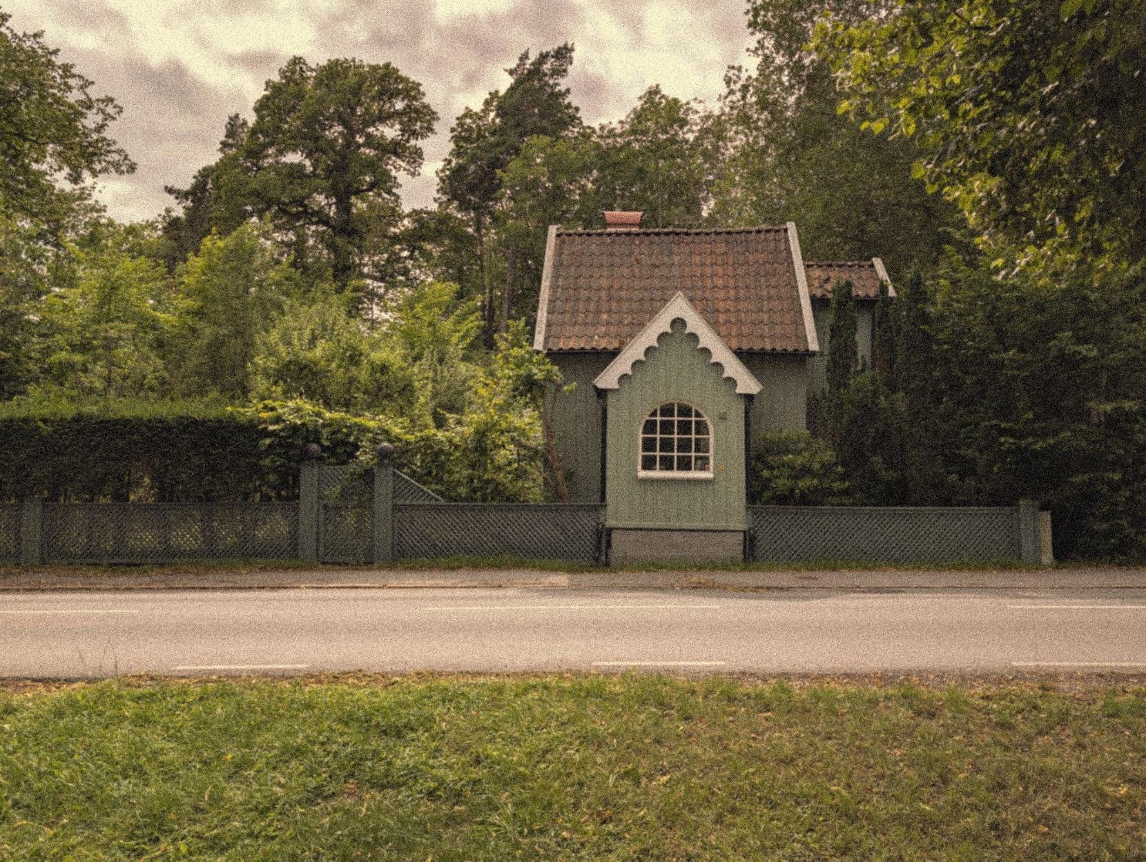 A grainy color photo of a small green house next to a road. One wall of the house is not behind the fence, but instead a part of it. Behind and next to the house are several lush green trees.