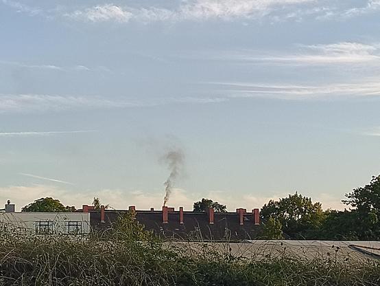 Vor blauem Himmel und Sonnenschein qualmt ein Schornstein eines Mehrfamilienhauses.
