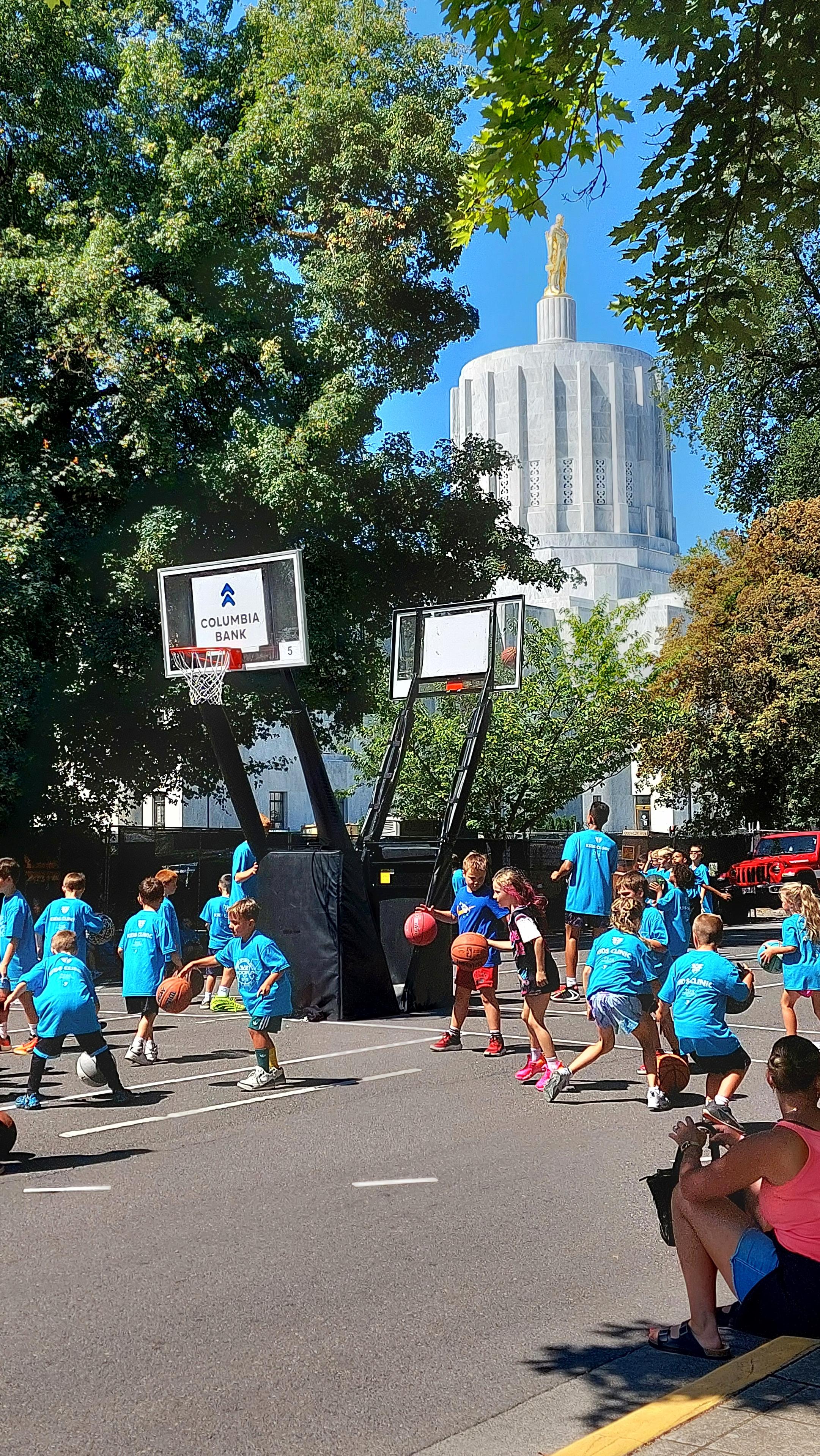 Children in aqua blue t shirts dribble basketballs under lowered baskets in front of the Oregon State Capitol building