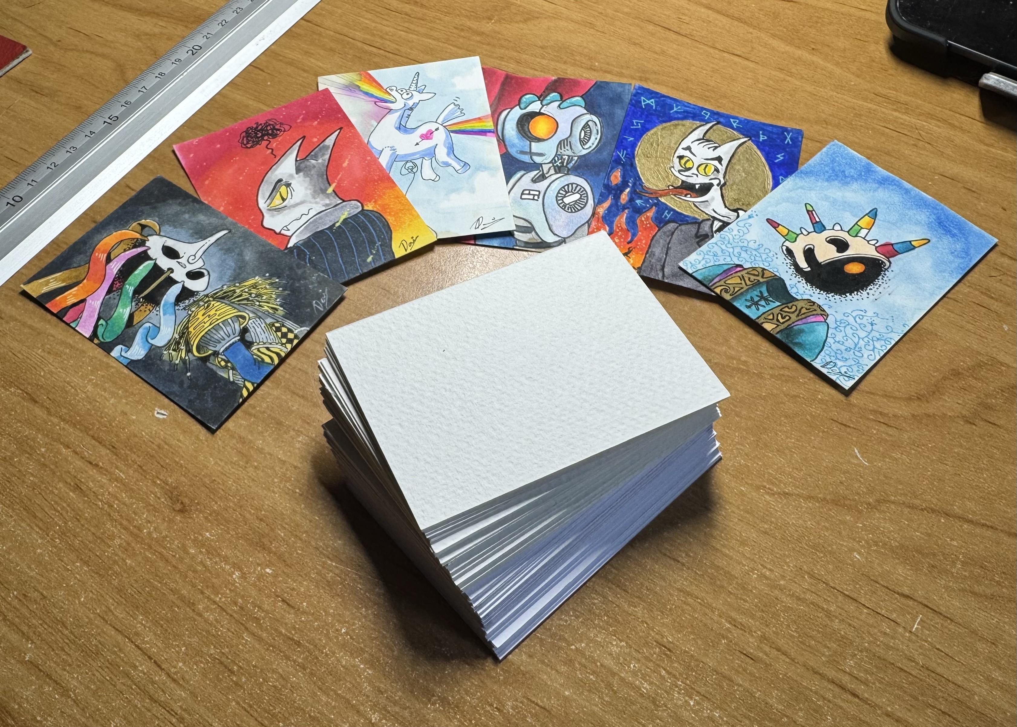 A stack of blank art cards is positioned on a wooden surface, surrounded by several colorful, illustrated cards featuring whimsical characters, including a unicorn, various animal-like figures, and a robot.