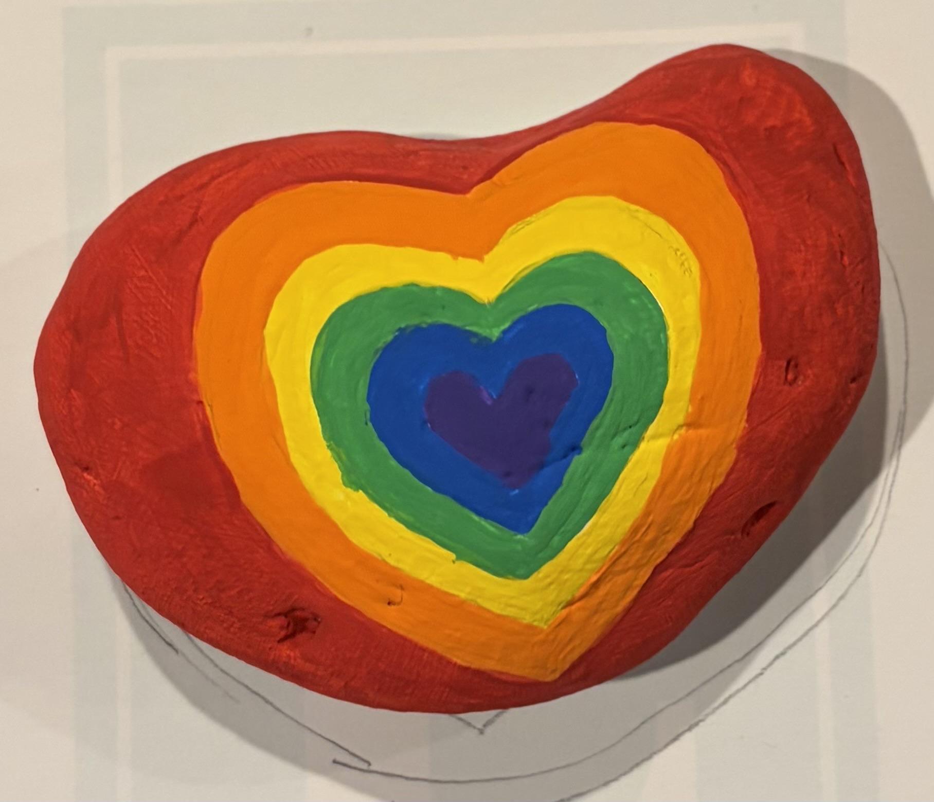 A hand-painted rock in the shape of a heart, featuring concentric rainbow-colored hearts (orange, yellow, green, blue, violet, from outside in) against a red background.