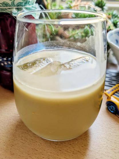 A close-up shot shows a clear, stemless glass filled with an opaque, light greenish liquid on a wooden surface. Two large ice cubes are partially visible near the top of the liquid inside the glass. In the blurred background, there is green foliage and a dark-colored object. To the right, part of a blue and white patterned bowl and a small yellow toy vehicle are visible.