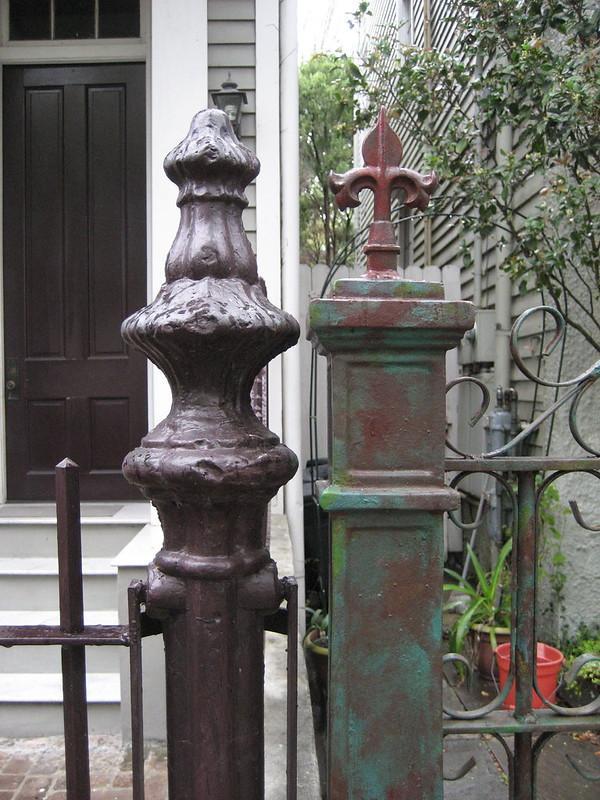 Two old metal fence posts side by side at a property line.  Small portion of wooden houses seen in background.
At left is a cylindrical vertical fence post painted dark brown, the top of which narrows then widens again in a vaguely plant-like pattern.  The visible bit of fence attached is a simple design of horizontal bars and vertical stakes in matching brown.
At right vertical rectangular fence post, the upper portion of which has a section with a square design topped with a fleur-de-lis.  Two layers of paint, dark red and green, are seen in uneven pattern - the post was no doubt painted solid colors at different times but parts of layers have worn away.  The visible bit of attached metal fence has series of decorative curls between and over the bars and stakes.