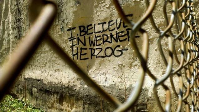 A close-up, slightly low-angle photo shot through a rusty chain-link fence in the foreground, which is partially out of focus. On the weathered, light-colored concrete wall behind the fence, black spray-painted capital letters spell out the phrase "I BELIEVE IN WERNER HERZOG". The wall shows signs of aging with peeling paint and dirt along the base.