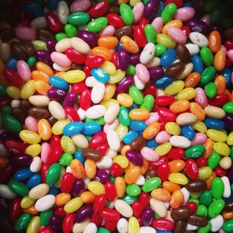 A colorful top-down, close-up photograph of a dense pile of jelly beans filling the entire frame. The candy piece collection features a vibrant mix of glossy colors, including bright red, pink, yellow, lime green, dark purple, orange, sky blue, white, and dark brown.