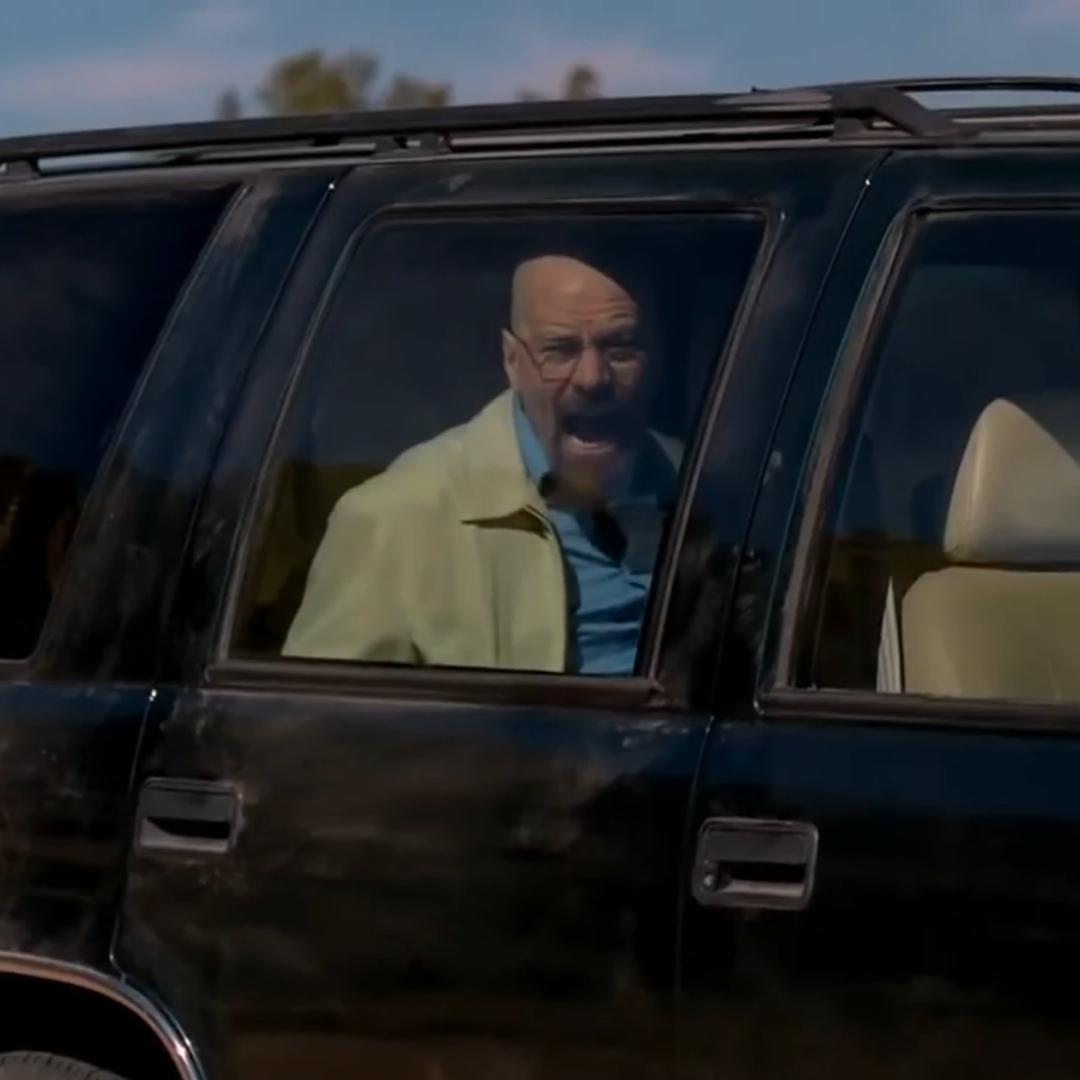 A man with glasses and a bald head shouts from the window of a dark SUV, surrounded by an outdoor setting.