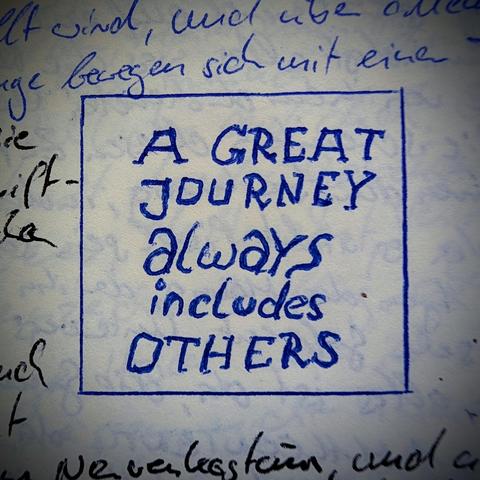 A handwritten note in blue ink on lined or textured paper, framed within a hand-drawn blue square. Inside the square, the text reads in capital and cursive-style letters: "A GREAT JOURNEY always includes OTHERS". Fragments of handwritten German text surround the framed square on the top, left, and bottom edges.