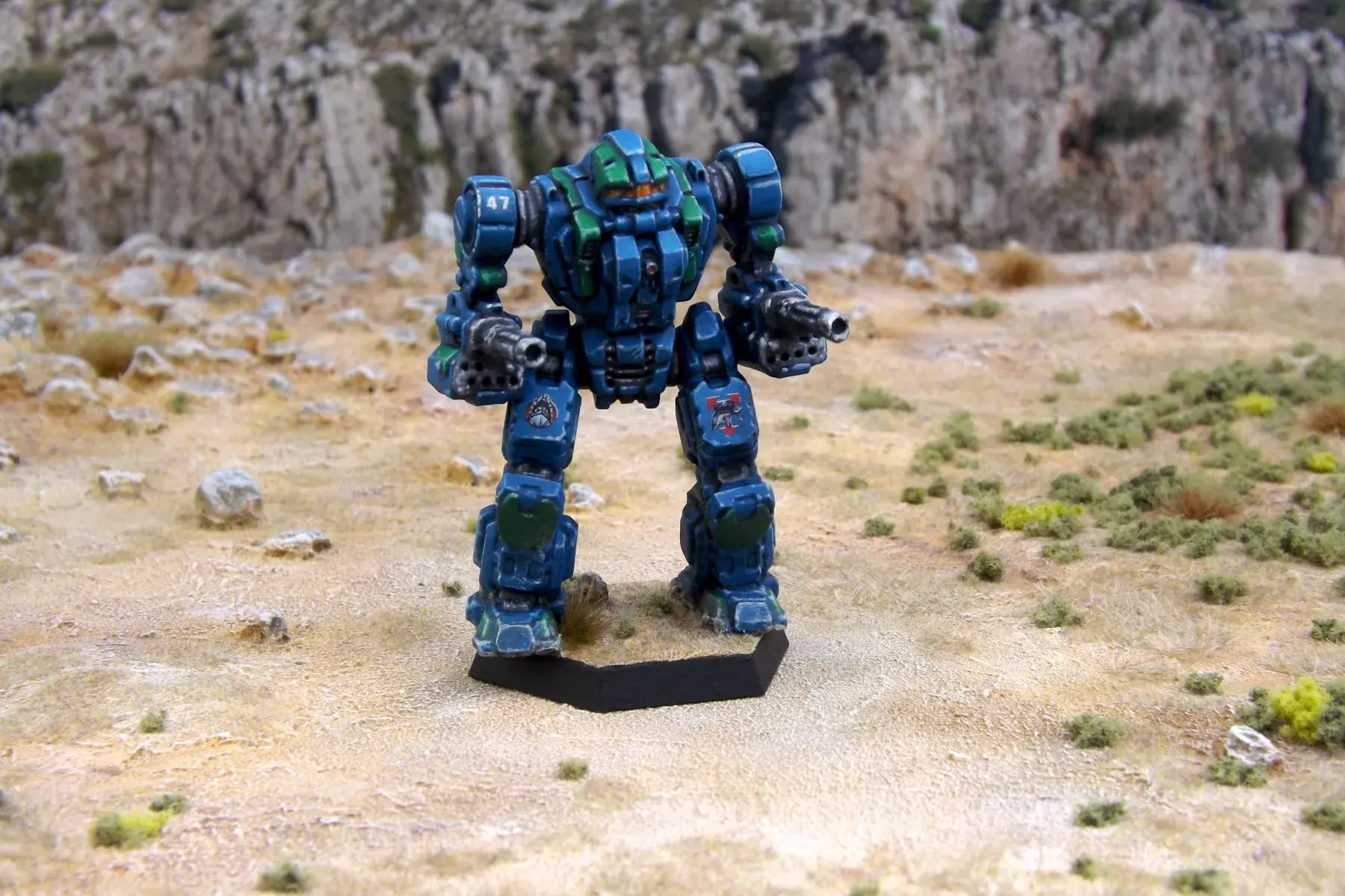 A painted miniature of a Gargoyle 'Mech.  It is blue with green highlights, and the cockpit glass is orange.  The number "47" is on the right shoulder.  The emblem of Clan Diamond Shark is on the left leg.  The emblem of Beta Galaxy, a shark's head over a red circle, is on the right leg.  The miniature is photographed on rocky terrain.