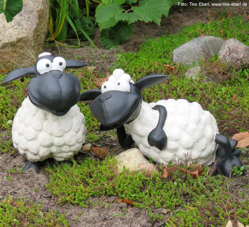 Zwei dekorative Schaf-Figuren mit übertrieben großen Augen und comicartigem Stil stehen auf einem Beet mit niedrigen grünen Pflanzen und kahler Erde. Die Figuren bestehen aus weißem, wollig strukturiertem Körper und schwarzen Gliedmaßen sowie Köpfen mit großen, runden Augen. Das linke Schaf steht aufrecht mit leicht geneigtem Kopf, während das rechte seitlich liegt und ein Bein nach vorne streckt. Im Hintergrund befinden sich größere Steine und grüne Blätter von Gartenpflanzen.