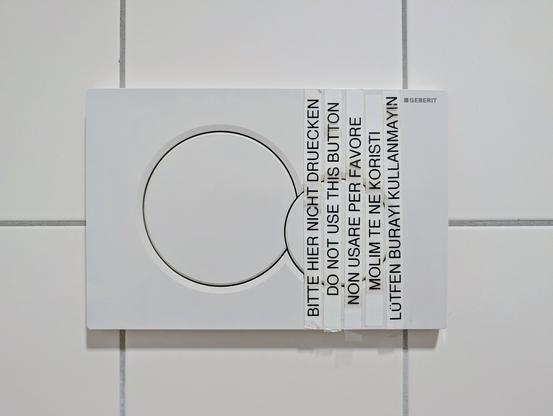 A white Geberit dual-flush toilet plate mounted on square tiled walls. Five label-maker tape strips are taped vertically across the right side and smaller flush button, displaying the message "do not use this button" in five languages:
 * German: BITTE HIER NICHT DRUECKEN
 * English: DO NOT USE THIS BUTTON
 * Italian: NON USARE PER FAVORE
 * Bosnian/Croatian/Serbian: MOLIM TE NE KORISTI
 * Turkish: LÜTFEN BURAYI KULLANMAYIN