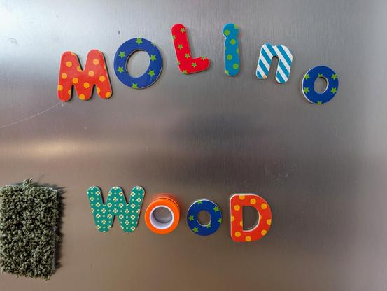 Colorful, patterned magnetic letters spell out the word "molino" above the word "WOOD" on a brushed stainless steel surface. The letter "o" in "WOOD" is an orange roll of tape. A fuzzy green magnet is on the far left.