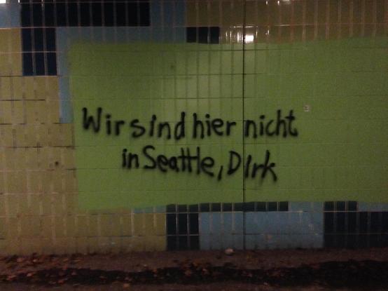 Foto einer gefliesten Wand im Außenbereich, vermutlich einer Unterführung. Auf einem großflächigen, hellgrünen Farbfeld, das ein älteres Mosaik oder Fliesenmuster überdeckt, steht in schwarzer Sprühfarbe in zwei Zeilen geschrieben: „Wir sind hier nicht in Seattle, Dirk“. Am unteren Bildrand ist ein Stück asphaltierter Weg mit einigen abgefallenen Blättern zu sehen.